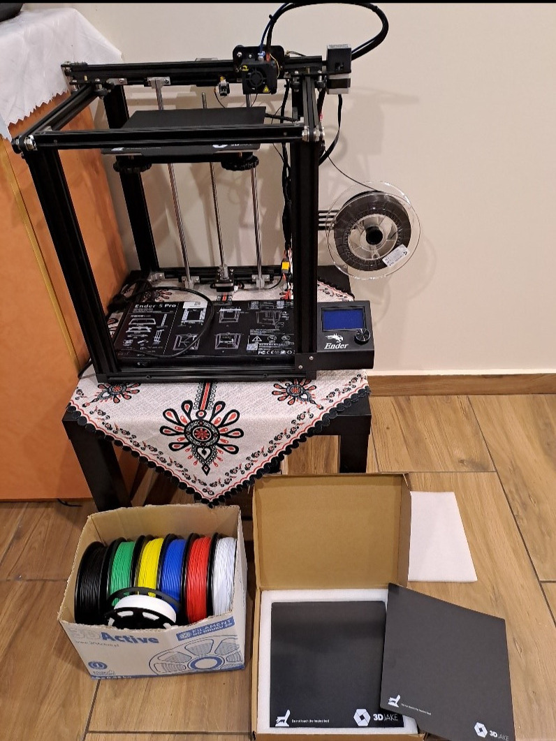 Drukarka 3D Ender S Pro na stoliku przy ścianie, obok pudełko z kolorowymi filamentami i akcesoriami 3DJake.