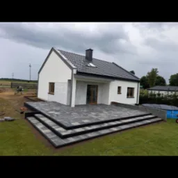 Nowoczesny dom jednorodzinny z tarasem z kostki brukowej w odcieniach szarości, schody z kostki brukowej prowadzą na trawnik, w tle widoczny basen i teren budowy.