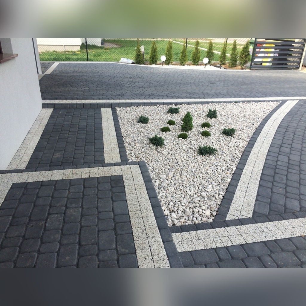 Ułożona kostka brukowa w kolorze grafitowym z jasnymi obrzeżami, tworząca ścieżkę i podjazd, z trójkątną rabatą wysypaną białym kamieniem i obsadzoną niskimi, zielonymi roślinami iglastymi.