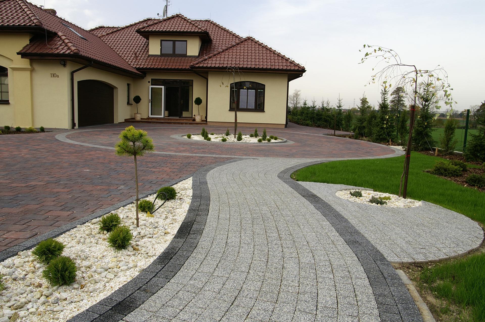 Elegancki podjazd do domu z kostki brukowej w odcieniach czerwieni i szarości, oddzielony pasem białych kamieni i niską zielenią, z widocznym budynkiem mieszkalnym w tle.