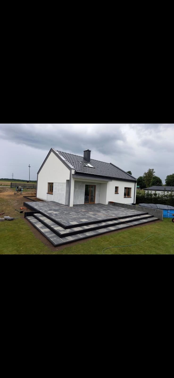 Nowoczesny dom jednorodzinny z tarasem z kostki brukowej w odcieniach szarości, schody z kostki brukowej prowadzą na trawnik, w tle widoczny basen i teren budowy.