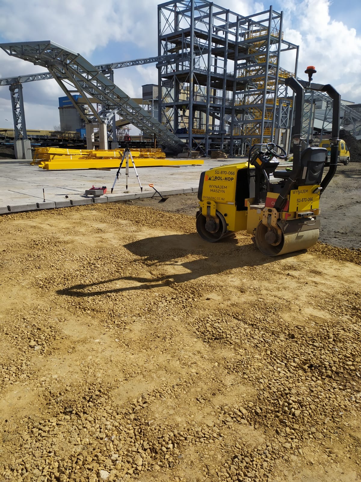 Żółty walec drogowy Dynapac CC 82 ubija żwir na placu budowy z konstrukcją stalową w tle, widoczny niwelator laserowy na statywie.