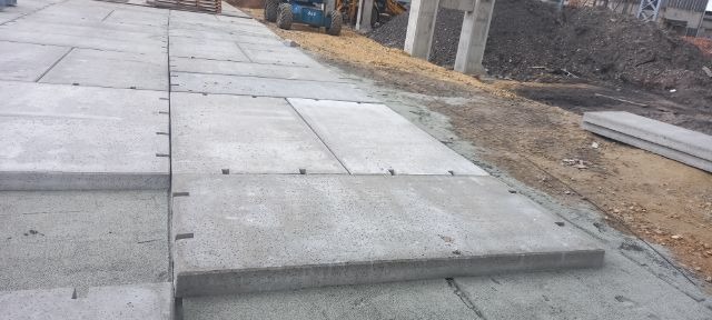 Układanie prefabrykowanych płyt betonowych na placu budowy, widoczne szczeliny dylatacyjne i ciężki sprzęt w tle.