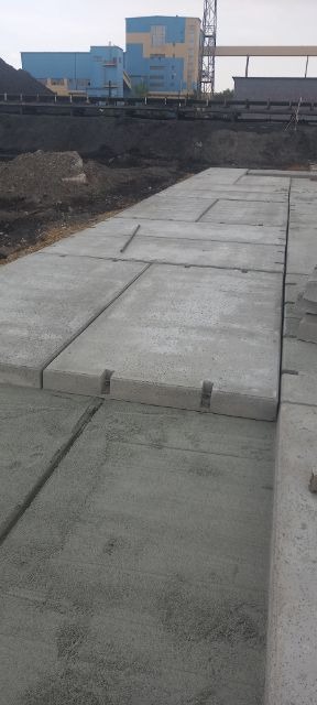 Ułożone, betonowe płyty chodnikowe z charakterystycznymi wycięciami w Pszczynie, tworzące utwardzoną powierzchnię na tle ziemi i budynków przemysłowych.