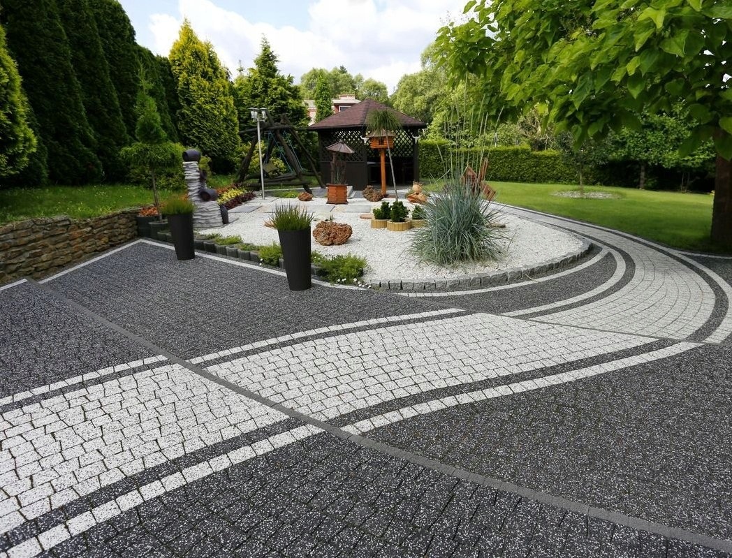 Ułożona kostka brukowa w odcieniach szarości i bieli, tworząca wzór z dekoracyjnym okrągłym klombem z białym żwirem i roślinnością ozdobną, w tle altana ogrodowa i zieleń.