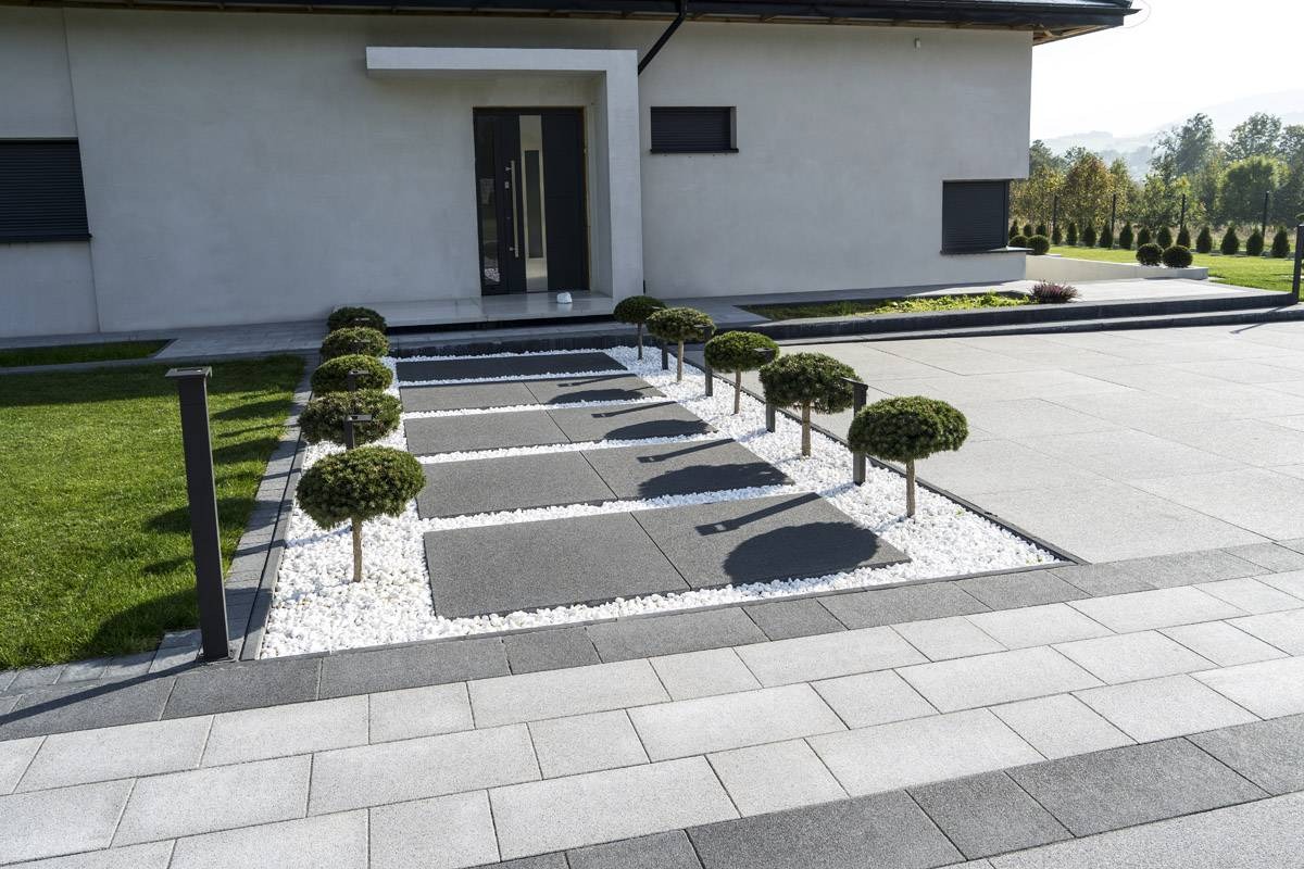 Nowoczesny podjazd z szarych płyt i białego grysu, ozdobiony kulistymi krzewami, prowadzący do minimalistycznego domu z ciemnymi drzwiami.