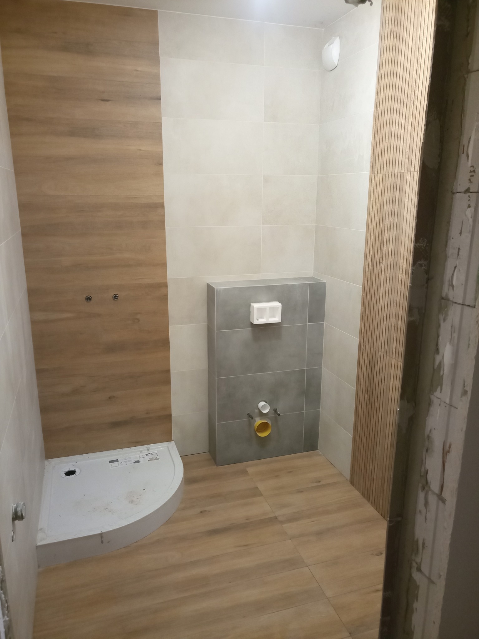 Wnętrze łazienki w trakcie prac: zamontowany brodzik, częściowo położone płytki imitujące drewno na podłodze i ścianie, szara zabudowa stelaża podtynkowego WC, widoczne przyłącza wodno-kanalizacyjne.