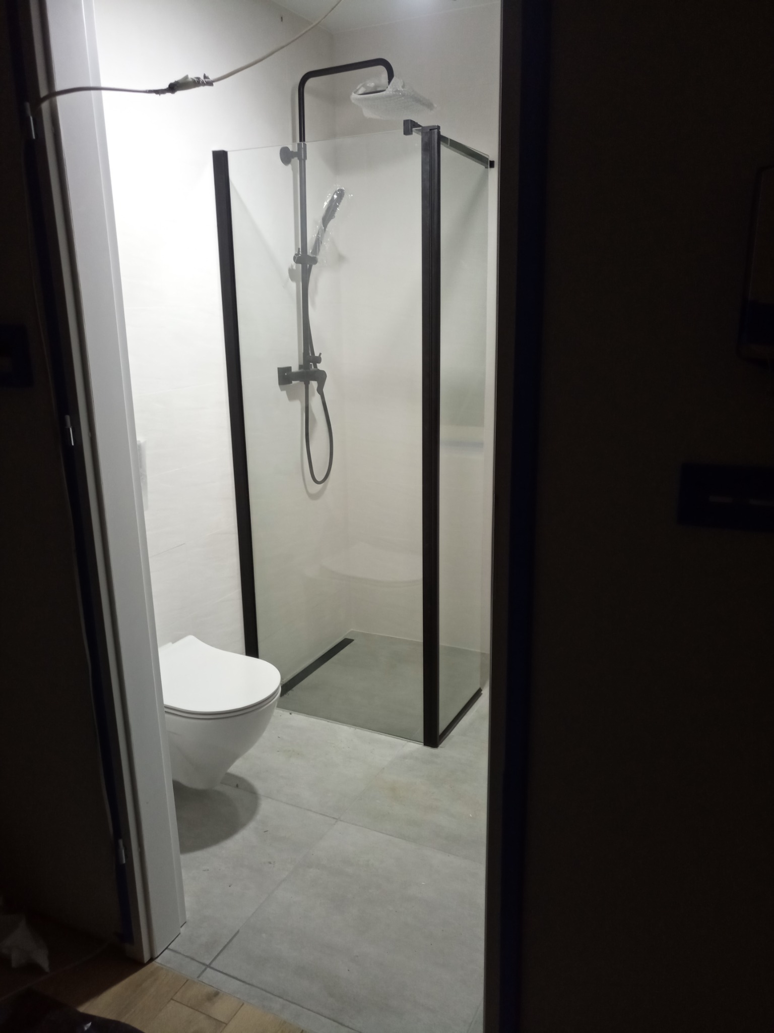 Nowoczesna kabina prysznicowa z czarnymi ramami i deszczownicą, widoczna toaleta i szare płytki podłogowe, ujęcie z perspektywy wejścia do łazienki.