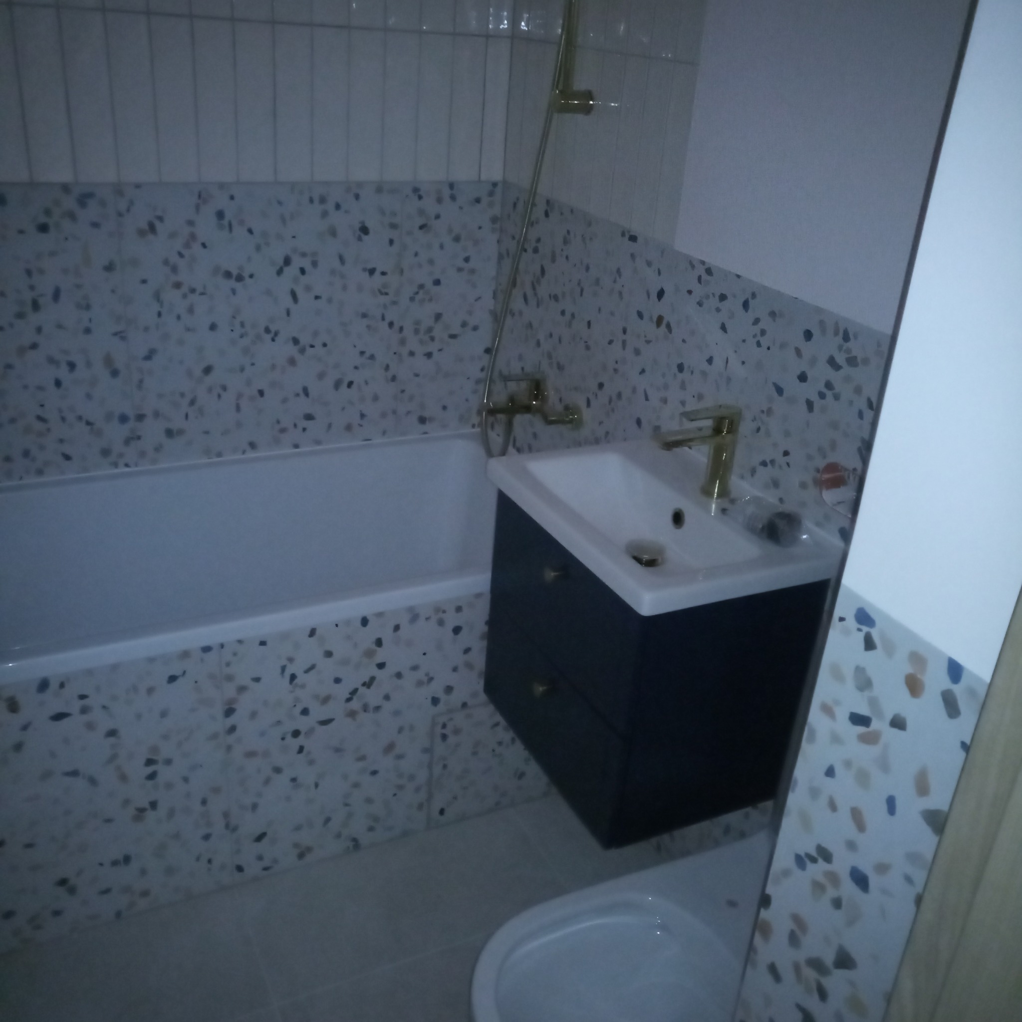 Wykończona łazienka z wanną, umywalką z szafką w kolorze granatowym, złotą armaturą i mozaikową glazurą na ścianach i obudowie wanny.