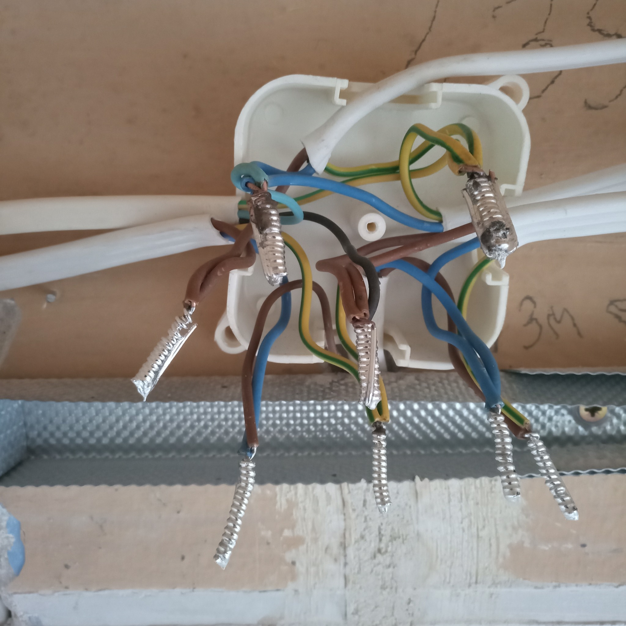 Połączenia elektryczne w puszce instalacyjnej z widocznymi przewodami w kolorach brązowym, niebieskim i zielono-żółtym, zabezpieczonymi srebrnymi tulejkami zaciskowymi, na tle sufitu podwieszanego...