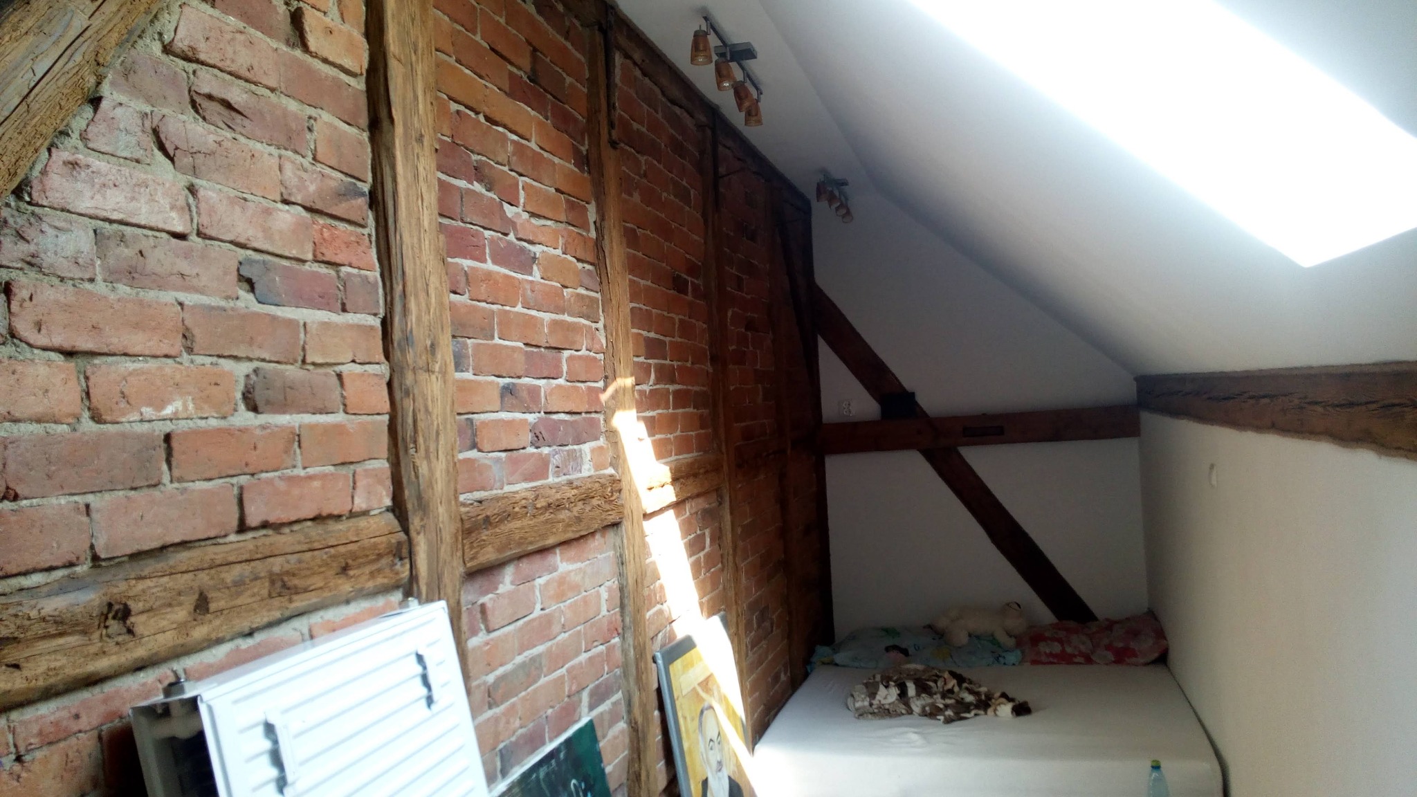 Pokój na poddaszu z odsłoniętą cegłą i drewnianymi belkami konstrukcyjnymi, oświetlony przez okno dachowe i lampy sufitowe.