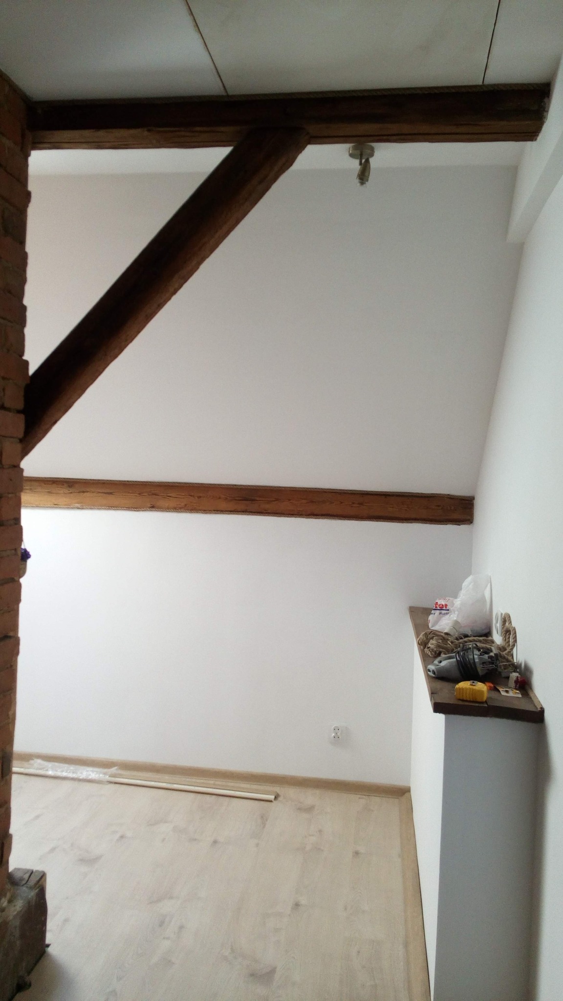 Wnętrze pokoju na poddaszu z drewnianymi belkami stropowymi, świeżo pomalowanymi białymi ścianami, jasną podłogą i narzędziami na półce.