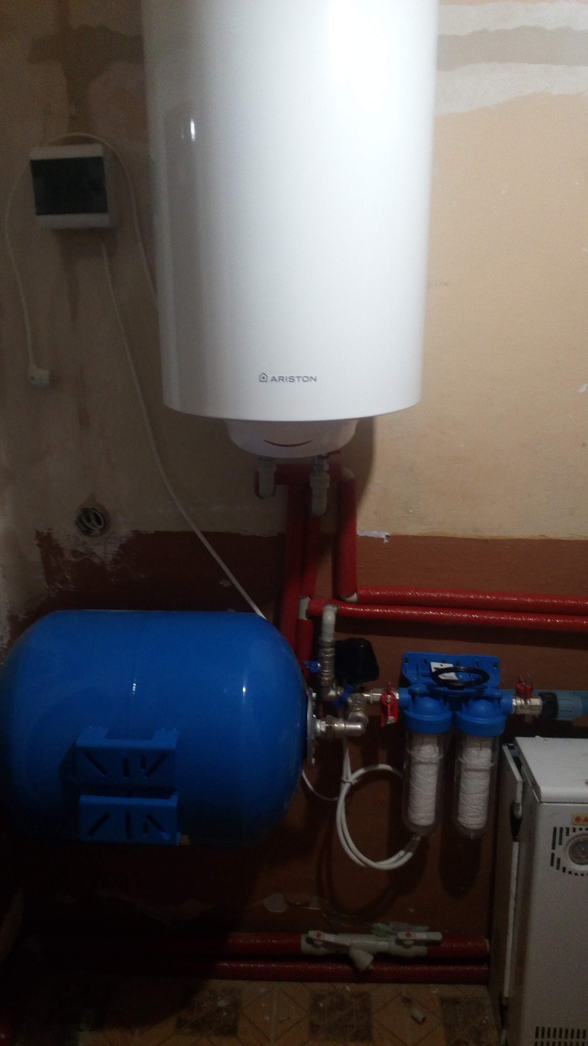 Bojler Ariston zamontowany na ścianie z widoczną instalacją hydrauliczną, filtrami wody i zbiornikiem wyrównawczym.