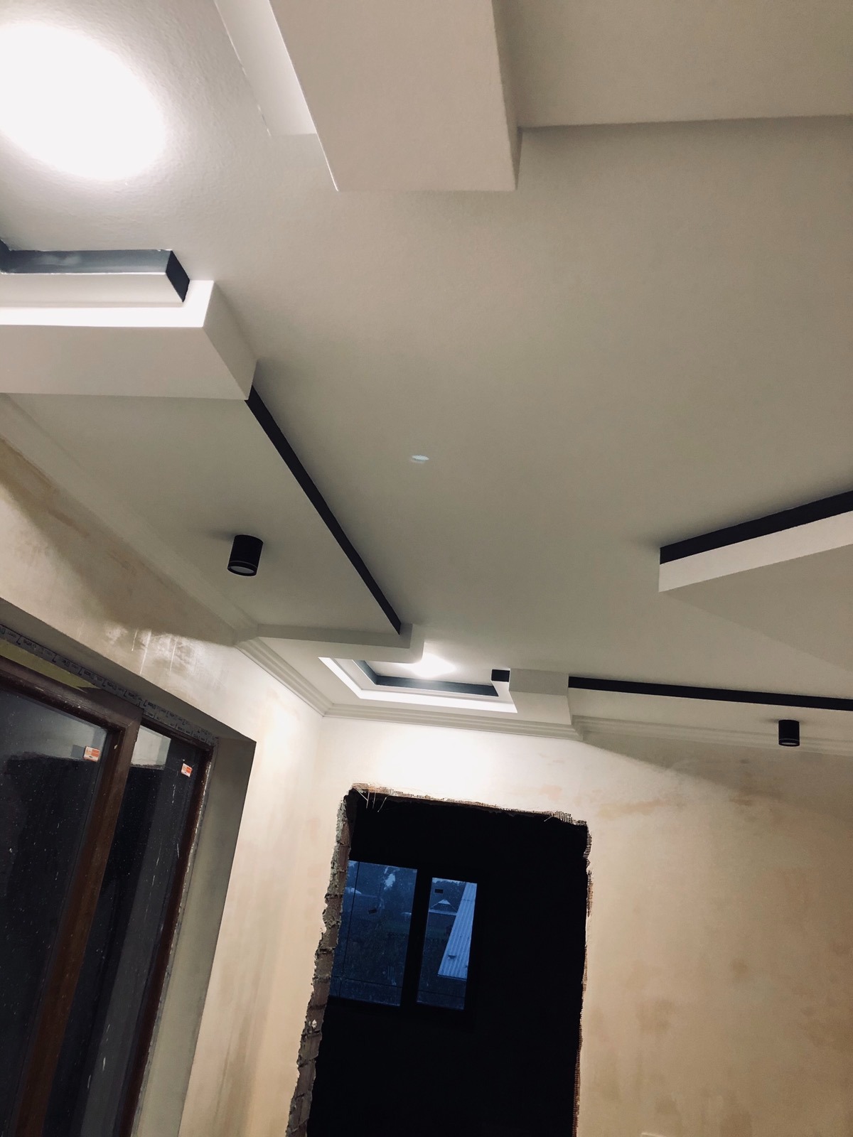 Wykończony sufit z podwieszanymi elementami dekoracyjnymi i oświetleniem punktowym, widok na niedokończone ściany i otwór drzwiowy.