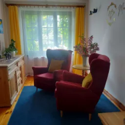 Przytulny gabinet z dwoma czerwonymi fotelami typu uszak, drewnianą komodą z dekoracjami, kwiatami w wazonie na małym stoliku i oknem z żółtymi zasłonami w tle.