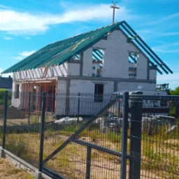 Wznoszenie dwukondygnacyjnego domu z betonu komórkowego H+H, widok zza czarnego ogrodzenia panelowego, widoczne elementy konstrukcji dachu i symbol budowlany na szczycie.