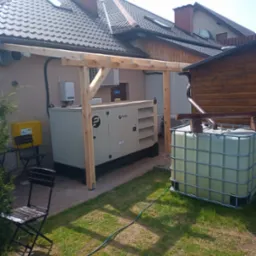 Agregat prądotwórczy przemysłowy marki Pezal umieszczony na zewnątrz budynku, z elementami konstrukcji z surowego drewna nad nim, obok zbiornik na deszczówkę, krzesła ogrodowe i żółta skrzynka gazowa.