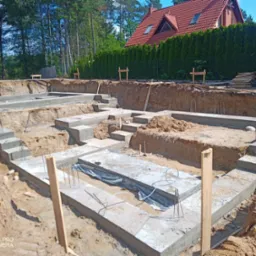 Zalane betonem fundamenty domu w wykopie, widoczne zbrojenia i drewniane szalunki, w tle zielony żywopłot i dom z czerwoną dachówką.