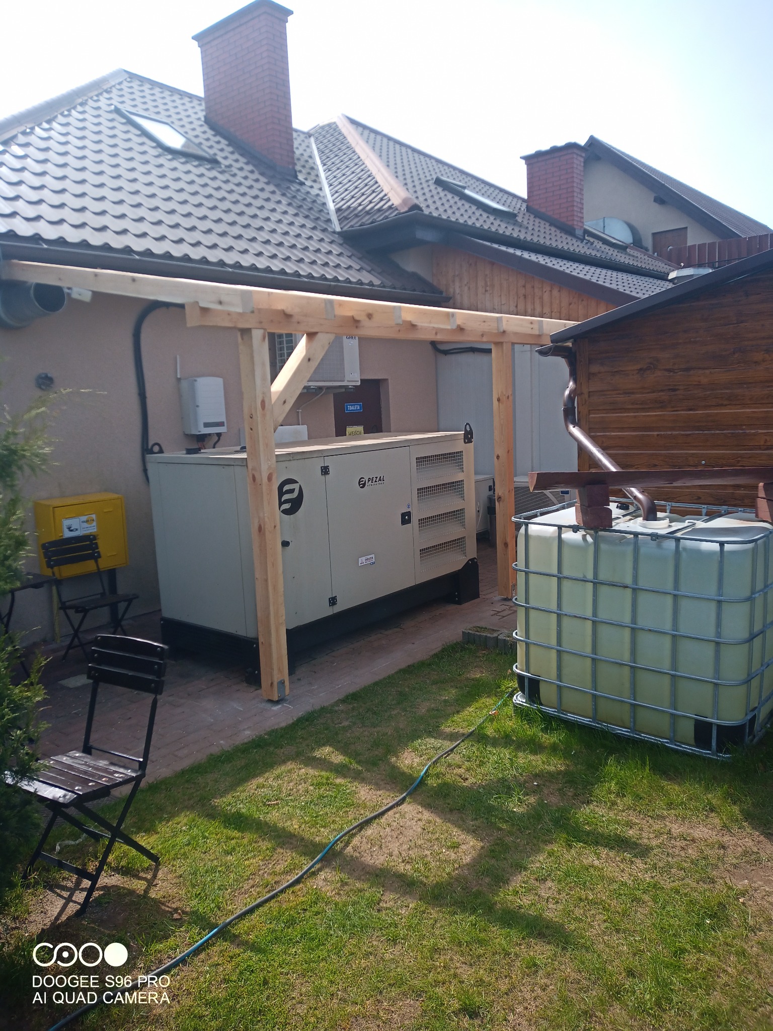 Agregat prądotwórczy przemysłowy marki Pezal umieszczony na zewnątrz budynku, z elementami konstrukcji z surowego drewna nad nim, obok zbiornik na deszczówkę, krzesła ogrodowe i żółta skrzynka gazowa.