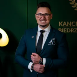 Uśmiechnięty prawnik w szmaragdowym garniturze, z przypinką orła, na tle zielonej ściany z logo kancelarii i lampą studyjną. Profesjonalny wizerunek.