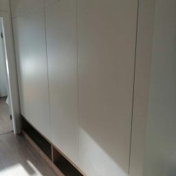 PioNik Meble na wymiar Piotr Nikiel - Biała szafa wnękowa z gładkimi frontami i otwartymi półkami u dołu, widoczna z boku na tle jasnej podłogi i ściany.