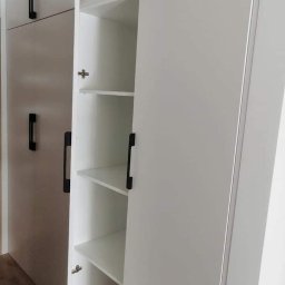 PioNik Meble na wymiar Piotr Nikiel - Wnętrze białej szafy z otwartymi drzwiami, widoczne półki i czarne, minimalistyczne uchwyty, sfotografowane z bliska.