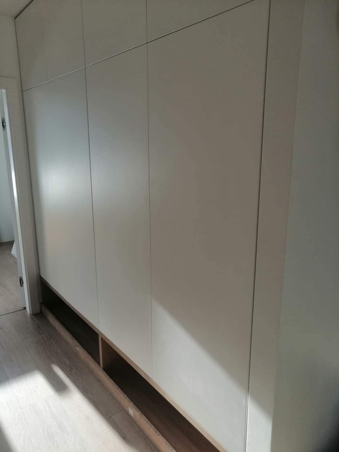 Biała szafa wnękowa z gładkimi frontami i otwartymi półkami u dołu, widoczna z boku na tle jasnej podłogi i ściany.