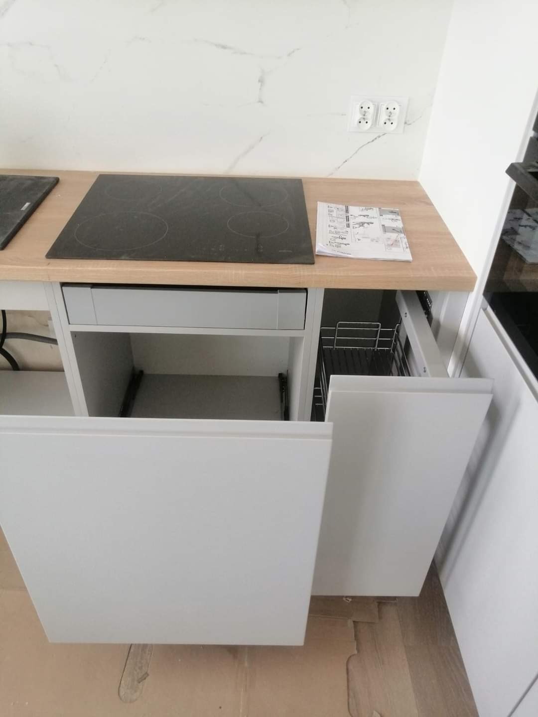 Fragment jasnej kuchni z otwartymi szafkami i szufladami, płyta indukcyjna na blacie drewnopodobnym, marmurowa ściana w tle.