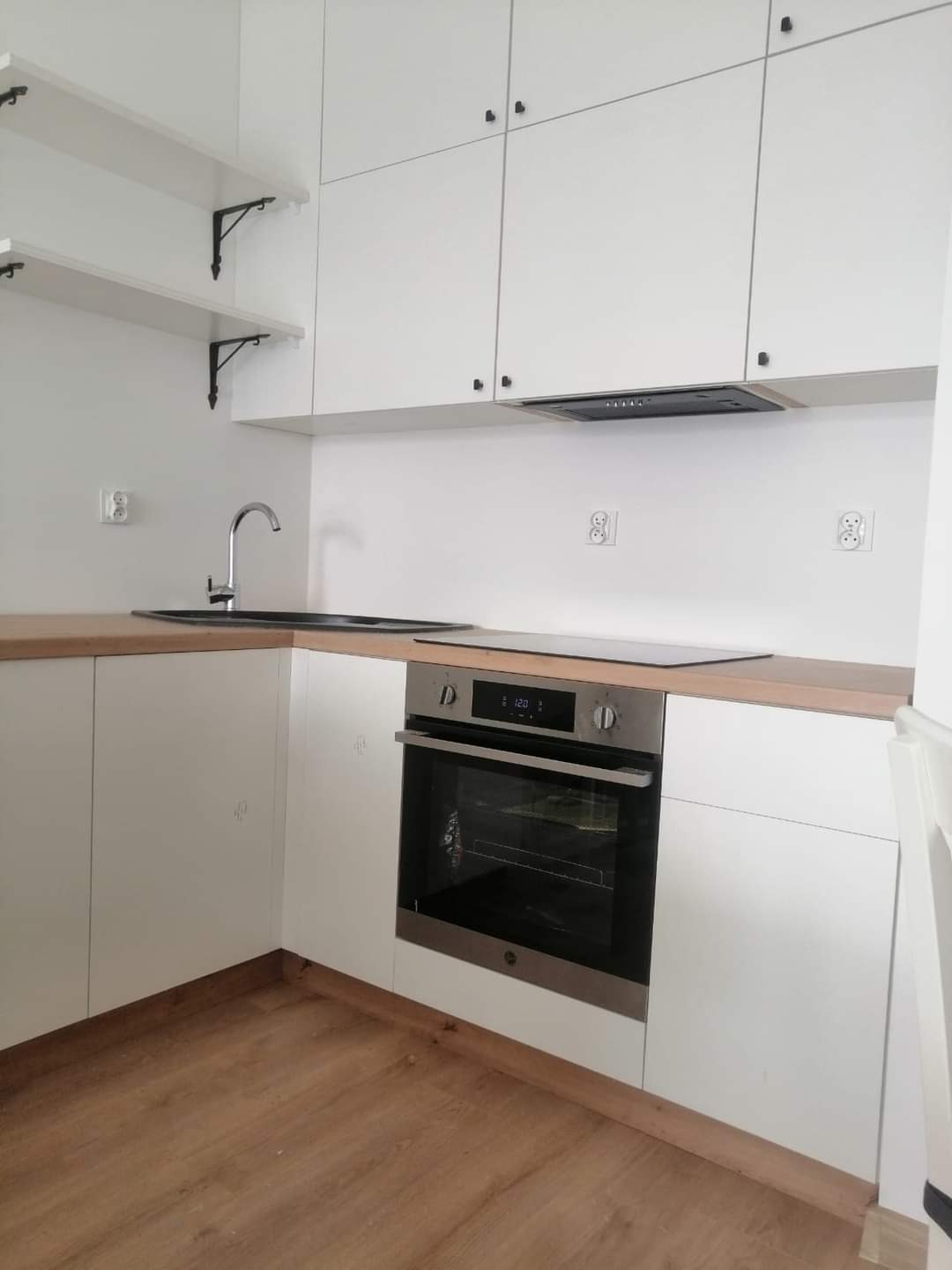 Minimalistyczna kuchnia: białe szafki z ciemnymi uchwytami, drewniany blat, piekarnik w zabudowie i otwarte półki z metalowymi wspornikami.