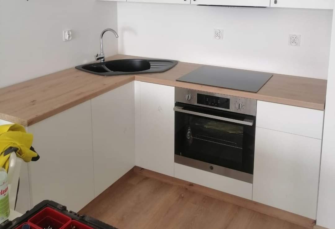 Fragment kuchni z drewnianym blatem, czarnym zlewem i płytą indukcyjną, widoczny fragment podłogi i narzędzi monterskich.