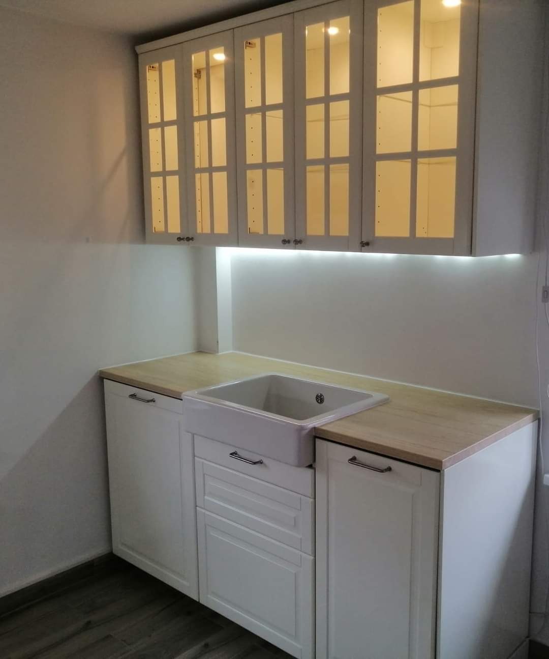 Biała kuchnia na wymiar z ceramicznym zlewem farmerskim i przeszklonymi szafkami górnymi z oświetleniem LED pod szafkami.