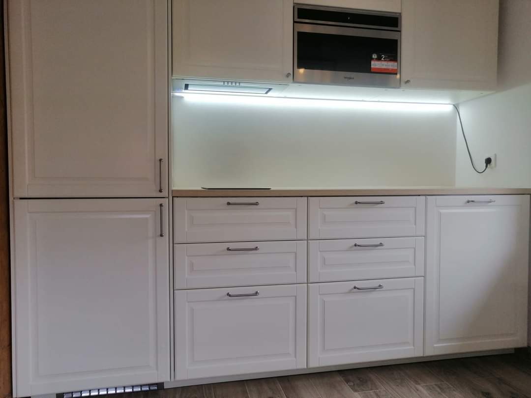 Klasyczna, biała zabudowa kuchenna z frezowanymi frontami, srebrnymi uchwytami i podświetleniem LED pod szafkami wiszącymi, widoczny fragment mikrofali w zabudowie.