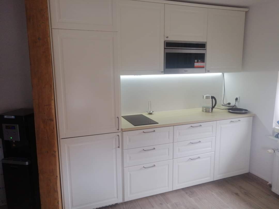 Jasna kuchnia z białymi szafkami, wbudowaną mikrofalówką i drewnianym blatem, oświetlona listwą LED, z widocznym dystrybutorem wody po lewej stronie