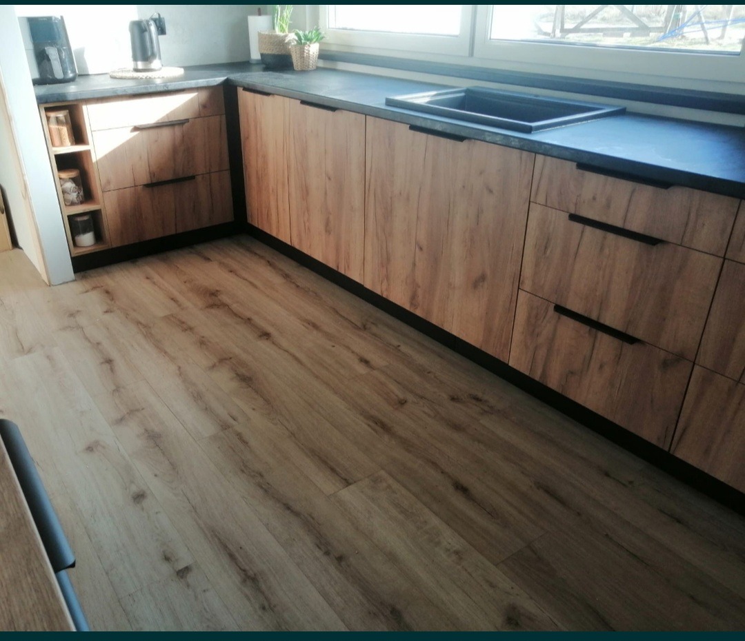 Kuchenna zabudowa z frontami w odcieniach drewna, czarnym blatem i minimalistycznymi uchwytami, uzupełniona o otwartą półkę na przyprawy i zioła przy zlewie.