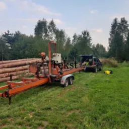 Przenośny trak taśmowy na przyczepie w trakcie pracy, obok stos drewna i samochód z otwartym bagażnikiem na trawiastym terenie.