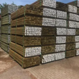 Równomiernie ułożone, impregnowane deski budowlane, zabezpieczone zielonymi taśmami, z widocznymi etykietami na końcach, składowane na zewnątrz na brukowanym placu.