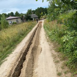 Marcel Wachnicki - Wykonane głębokie wykopy w piaszczystej ziemi na wiejskiej drodze, widoczne ślady opon, w tle dom i zieleń.