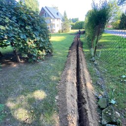 Marcel Wachnicki - Długi, wąski wykop w ziemi na trawniku wzdłuż siatki ogrodzeniowej, w tle dom jednorodzinny i pracująca osoba.