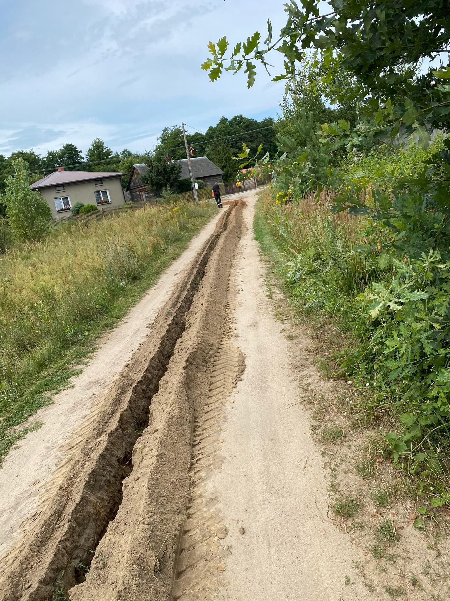 Wykonane głębokie wykopy w piaszczystej ziemi na wiejskiej drodze, widoczne ślady opon, w tle dom i zieleń.