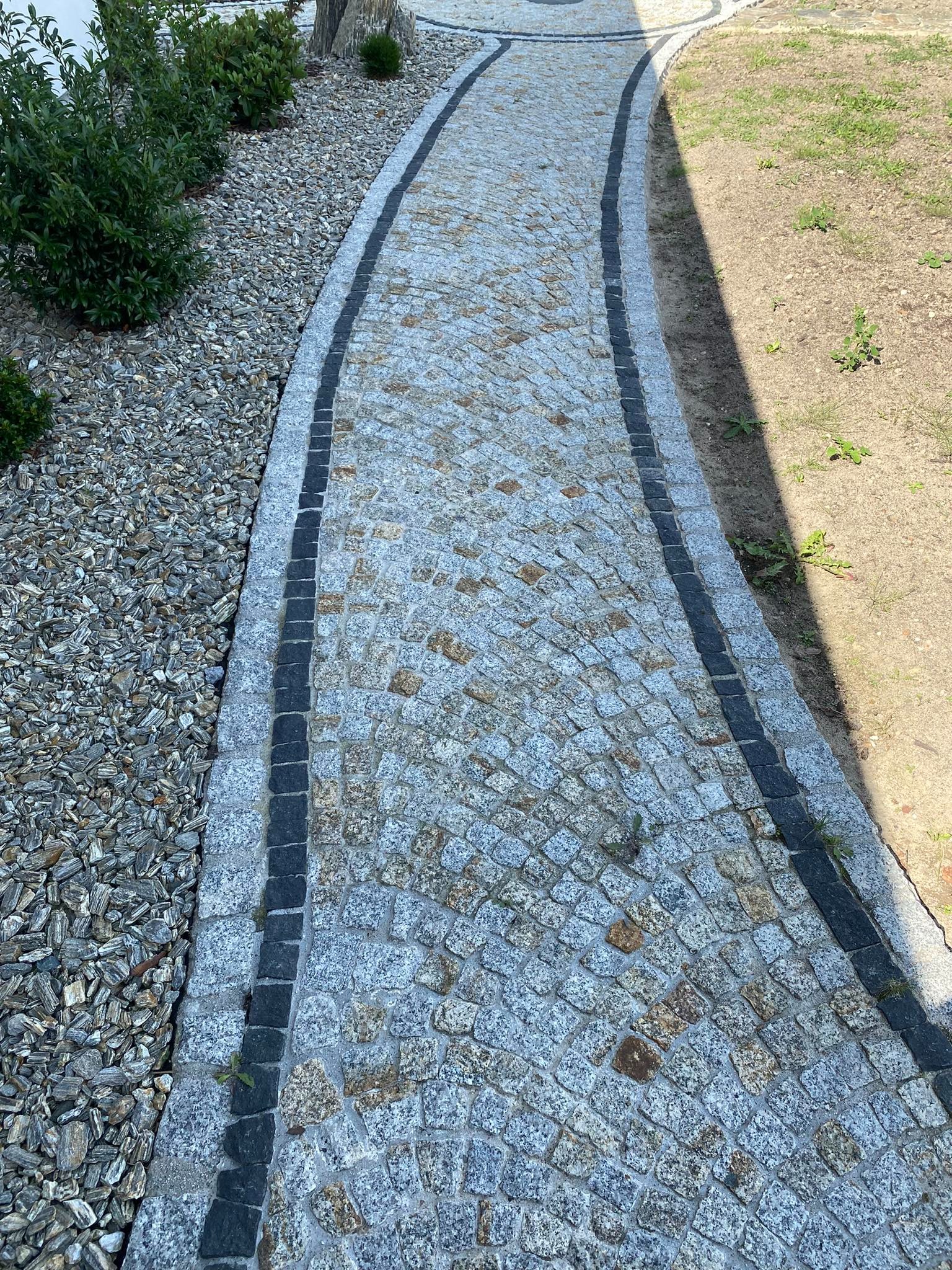 Zakrzywiona ścieżka z kostki granitowej w odcieniach szarości i brązu, obrzeżona ciemnoszarą kostką, przylegająca do kory drzewnej i trawnika z rzadką roślinnością.