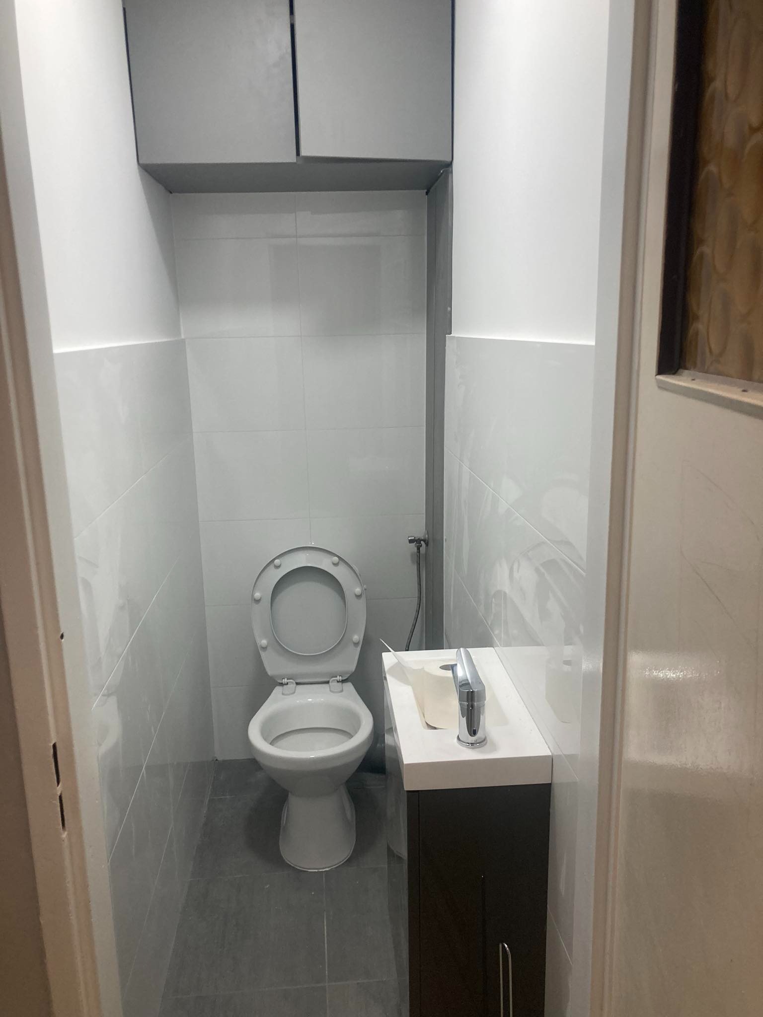 Wąska łazienka po remoncie z szarymi płytkami, podwieszaną toaletą, szafką z umywalką i baterią, oraz szafkami wiszącymi pod sufitem.