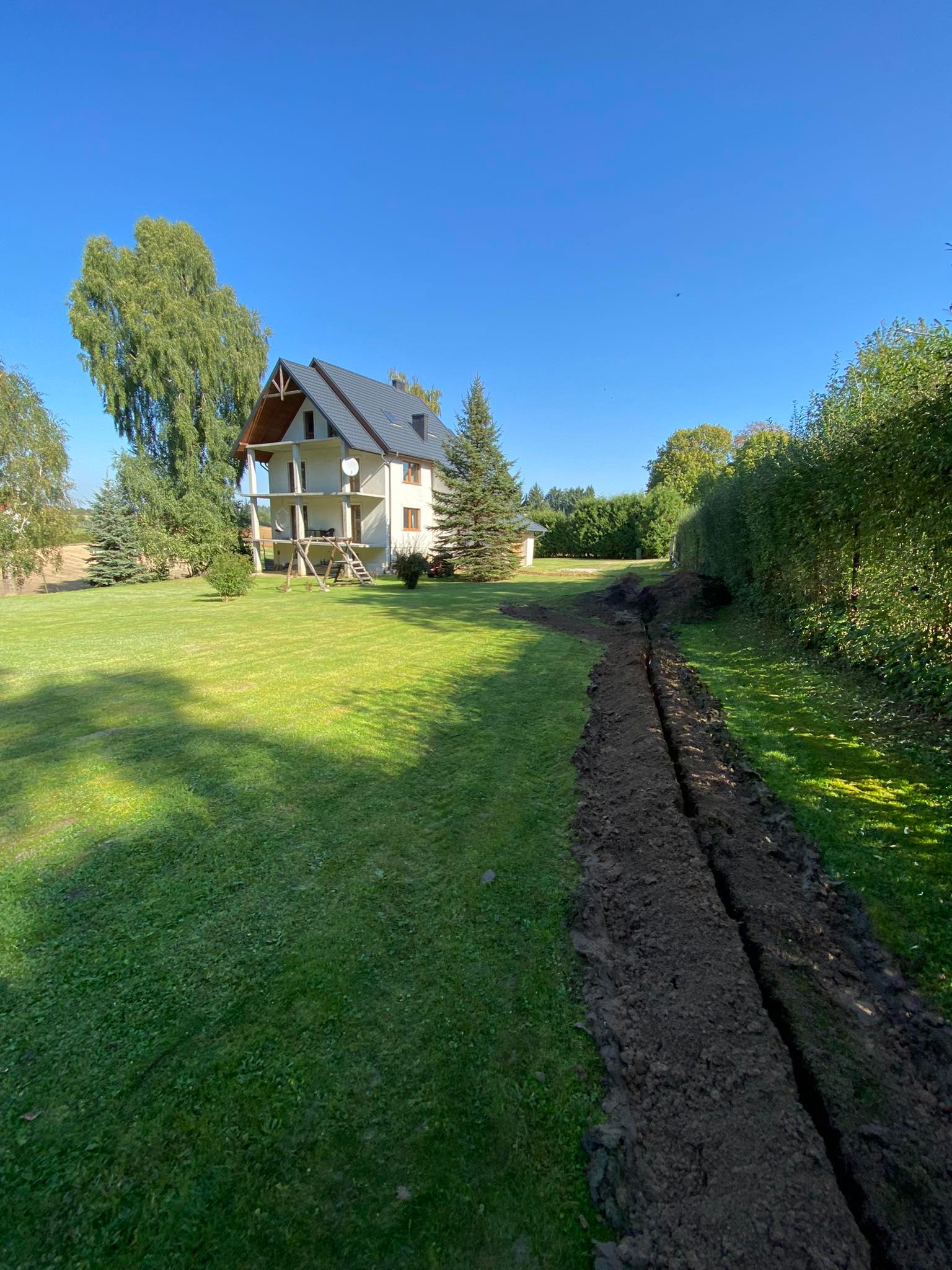 Wykopany rów w trawniku na posesji z domem jednorodzinnym w tle, widoczne ślady świeżej ziemi.