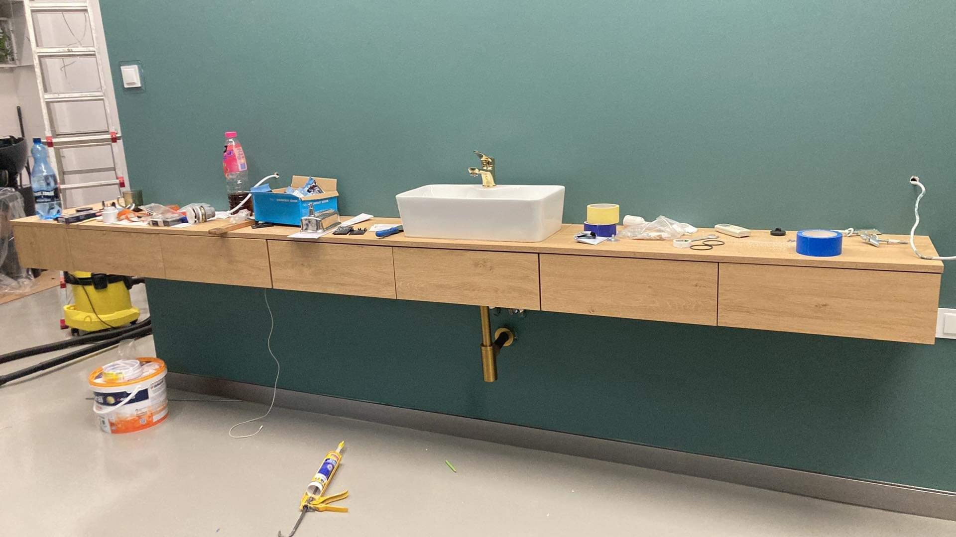 Montaż wiszącej szafki łazienkowej z umywalką nablatową i złotą armaturą, widoczne narzędzia i akcesoria montażowe na blacie, zielona ściana w tle, etap prac wykończeniowych.