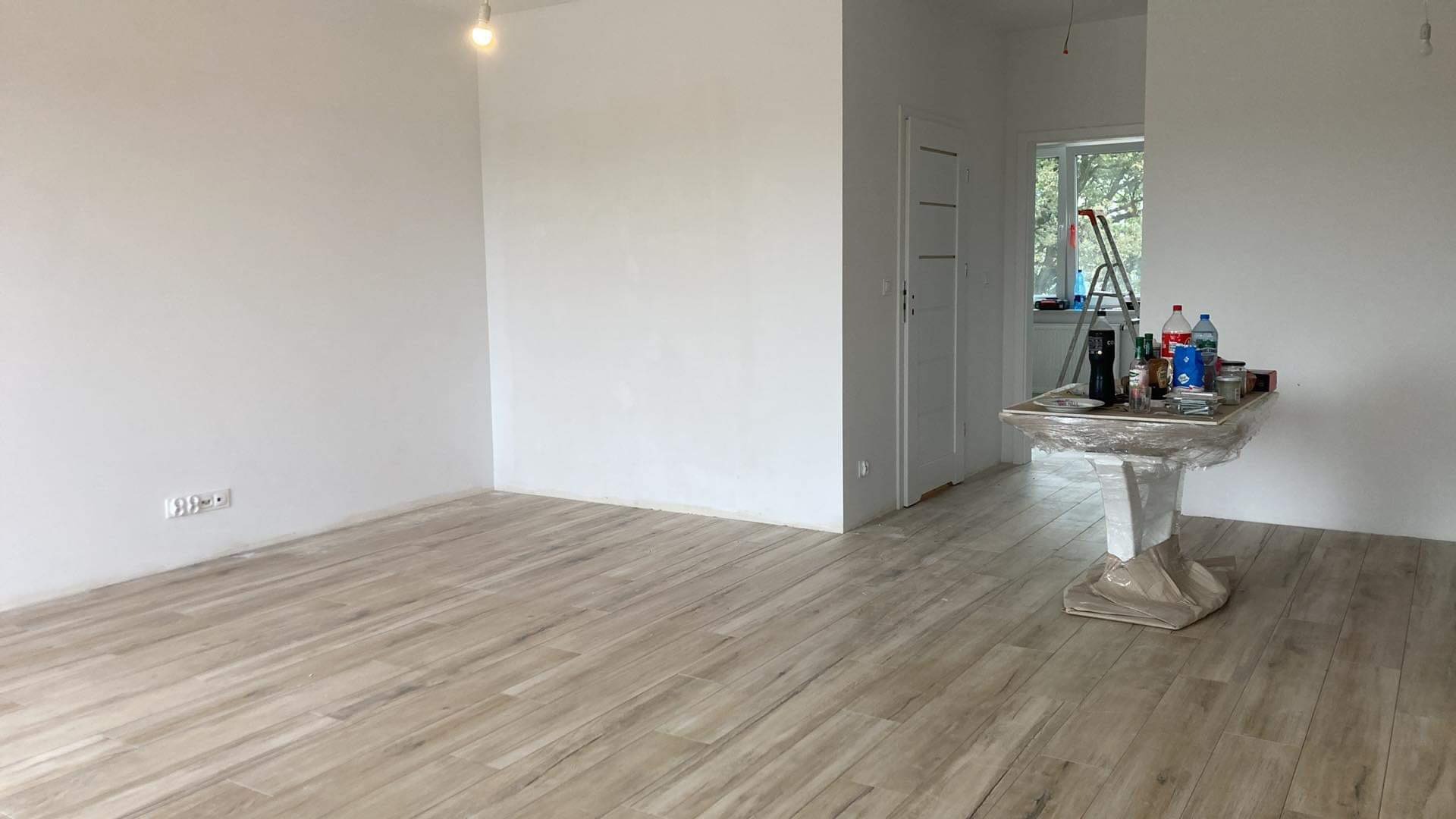 Wnętrze pokoju z ułożoną podłogą z paneli imitujących drewno, świeżo pomalowanymi ścianami na biało, osprzętem malarskim na stole zabezpieczonym folią oraz drabiną w tle przy oknie.