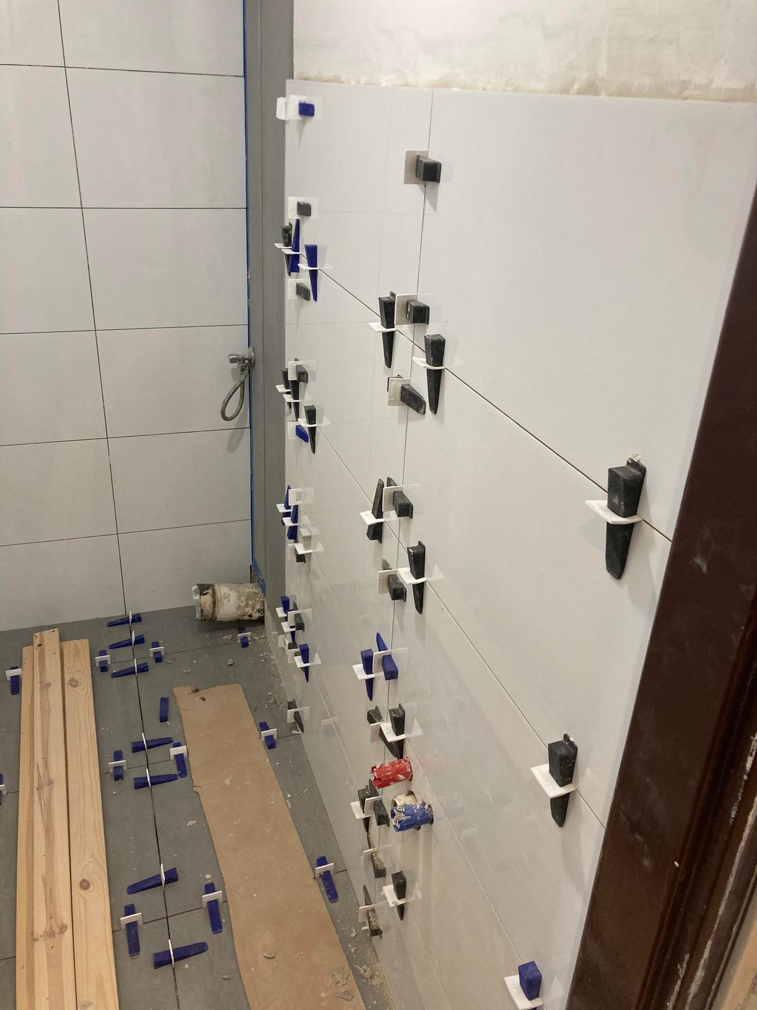 Ściana łazienki w trakcie układania białych płytek z użyciem klipsów poziomujących i krzyżyków dystansowych, widoczne rury i deski na podłodze.