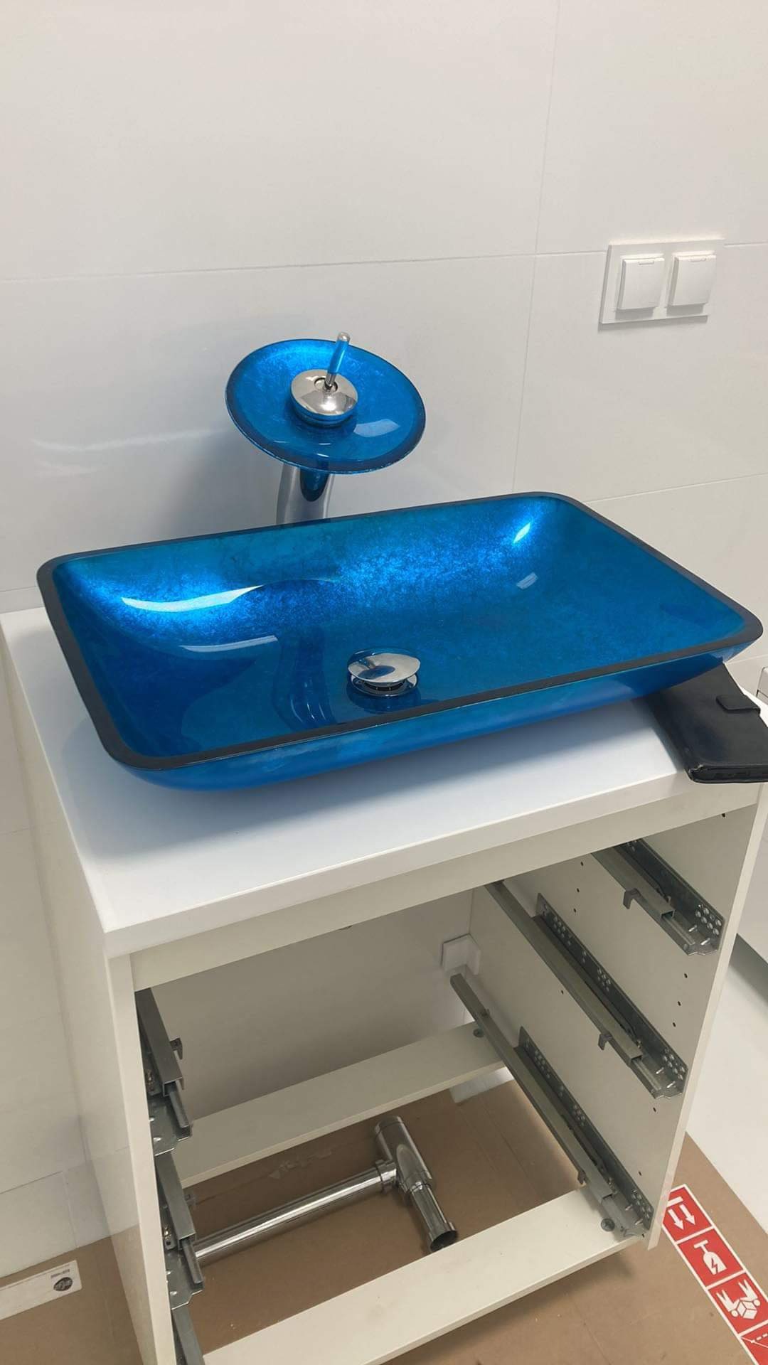 Błękitna, szklana umywalka nablatowa z chromowaną armaturą na białej szafce, widoczne elementy montażowe szuflad i syfonu, białe płytki na ścianie.