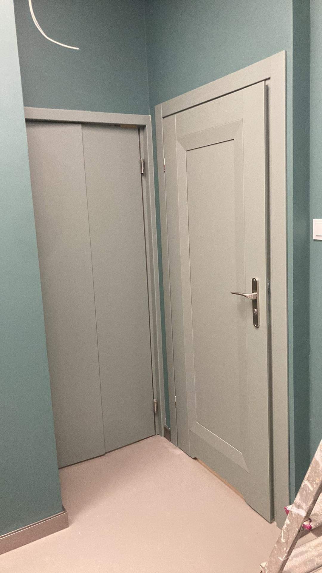 Dwie pomalowane na szaro drzwi wewnętrzne w nowym mieszkaniu, widoczna drabina malarska w rogu, ściany w odcieniu turkusowym.