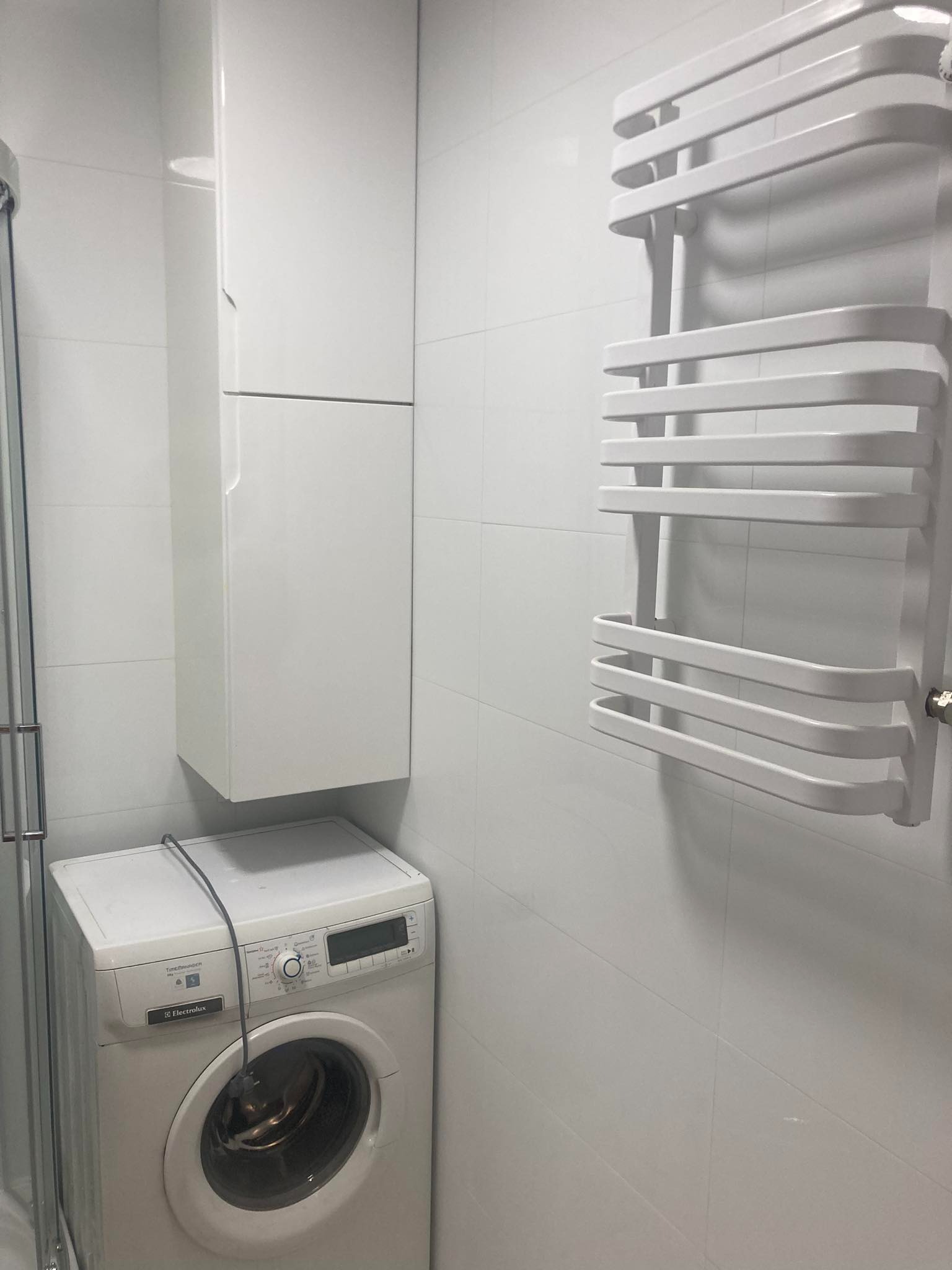 Biała łazienka z pralką Electrolux, wiszącą szafką i grzejnikiem drabinkowym na tle białych płytek