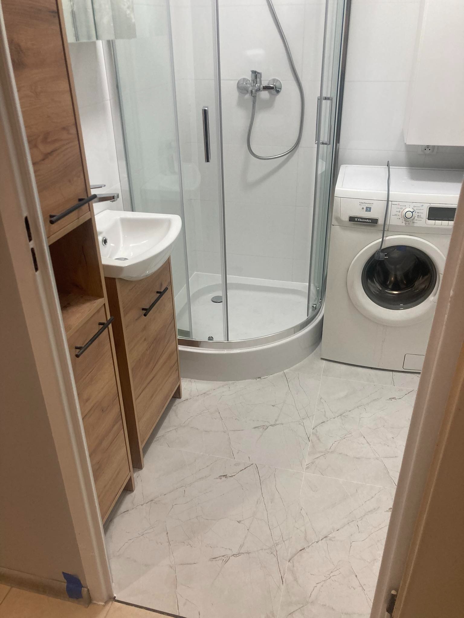 Wykończona łazienka z kabiną prysznicową, umywalką w zabudowie z drewnopodobnymi szafkami i pralką Electrolux na marmurowej posadzce.