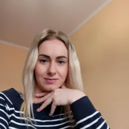 Portret kobiety z blond włosami w bluzie w paski, opierającej brodę na dłoni, z delikatnym uśmiechem, na tle beżowej ściany i białego sufitu.